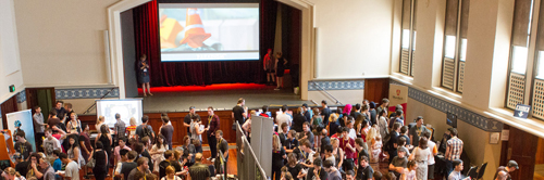 Banner: The Perth Town Hall, filled with people crowding around various game screens and tables. A screen hangs above the stage, showing the image of local game 'Paradigm'