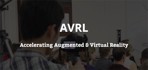 Image text reads: "AVRL. Accelerating Augmented & Virtual Reality". Darkened background image depicts a group of people from behind, looking towards a whiteboard. The man in the foreground is holding up a camera to record the presentation they are watching.