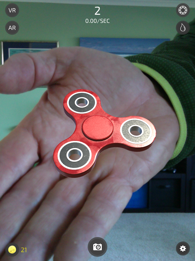 A screenshot from the game Fidget Spinner: Journey of Secrets. The screenshot shows a photograph of a man's open hand with the palm facing the viewer. In this hand sits a 3D fidget spinner, the model overlayed using Augmented Reality. Around the edges of the screen are the user interface for the game.