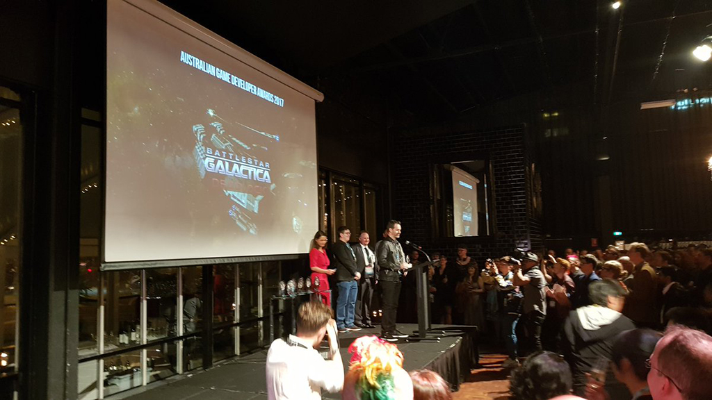 A photograph from the AGDA event,; Paul Turbett and the Black Lab Games team receives their award, as the screen behind them displays the game title .