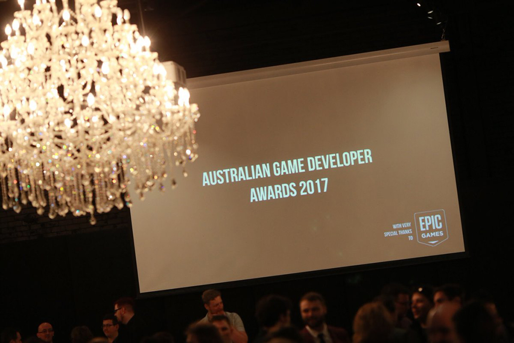 A projector screen sits above a crowd of people, framed to the left by a chandelier. On the screen, text is displayed which reads "Australian Game Developer Awards 2017",