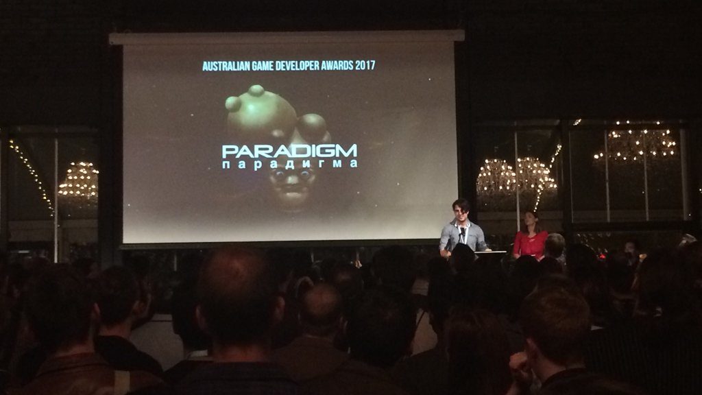 A photograph from the AGDA event,; Jacob Janerka receives his award, as the screen behind him displays the game title Paradigm and its main character.
