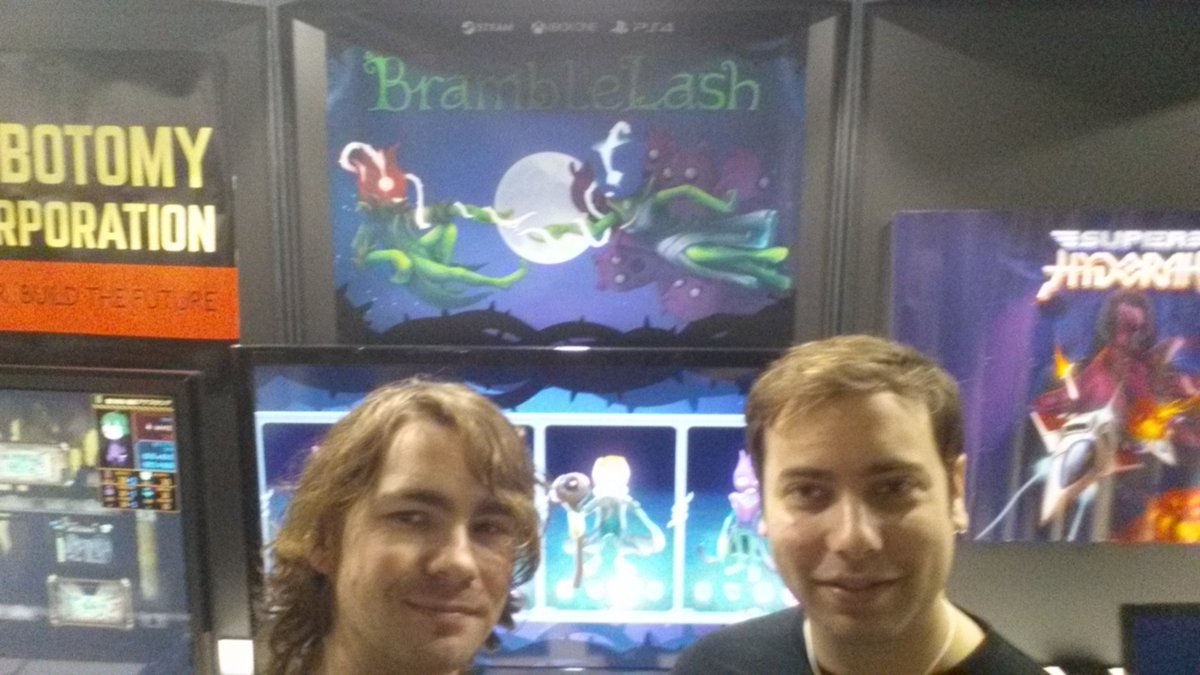 The team members from Bytesprite, Liam and Sam, pose for a photograph in front of the BrambleLash booth. In the background the game's character selection screen is being displayed on a monitor, and the promotional poster hangs on the back wall.