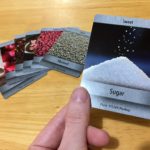 A hand hold up the Sweet/Sugar ingredient card. On the table a collection of other food-based cards are laid out.