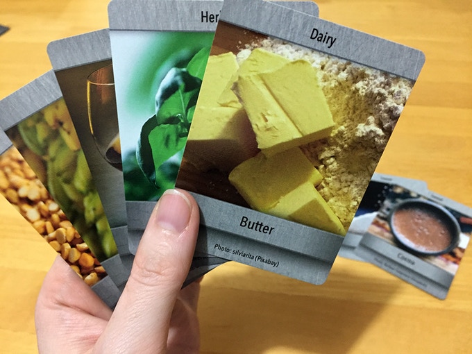 A hand hold up a selection of game cards, with the Dairy/Butter ingredient card at the top. On the table a collection of other food-based cards are laid out.