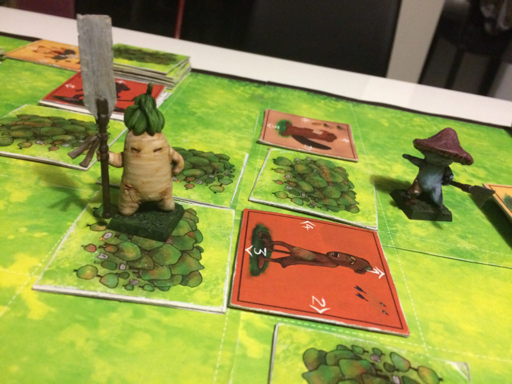 Photograph: A tabletop game with a board depicting grass and trees, and two large models of game characters sitting upon it. One, a radish with short arms and legs holds a long blunted spear weapon, the other, a smaller Mushroom-like person pointing an also brandishing a similar weapon.