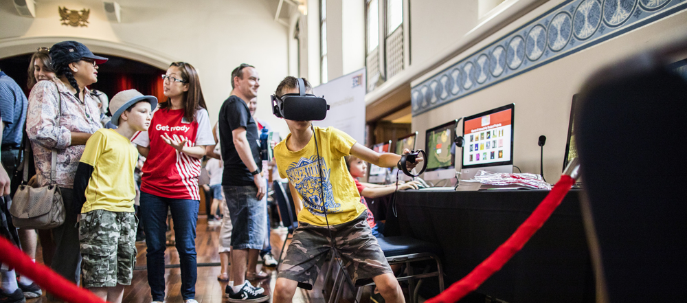 A young boy in the foreground wears a virtual reality headset and is punching his arm forwards. In the background is a crowd of people playing computer games at the Perth Town Hall.