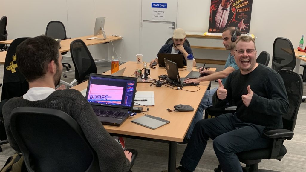 Photograph of people with laptops out working at a table. One man smiles at the camera and giving two thumbs up.