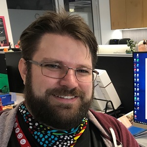 Photograph of Matt Dyet, LMG Chairperson as of 2019. A caucasian man smiling with short brown hair, beard and moustache. He has thin metal glasses, and wears a bandanna around his neck.