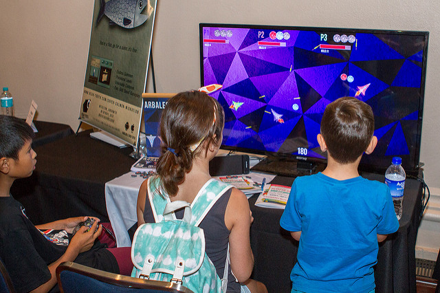3 kids sit in front of a large screen, playing a stylised spaceship game together.