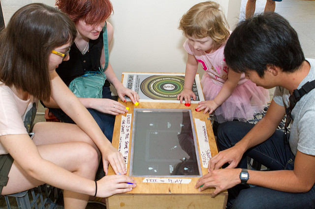 3 young adults and a small child sit around a table that has a digital game built into it's surface.