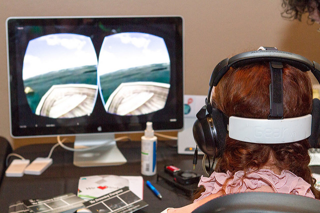A woman wears a virtual reality headset, and a view from within the game can be seen on the screen in front of her.